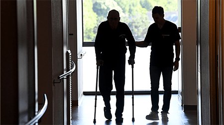 Dans une clinique, un soignant aide une personne âgée à marcher avec des béquilles
